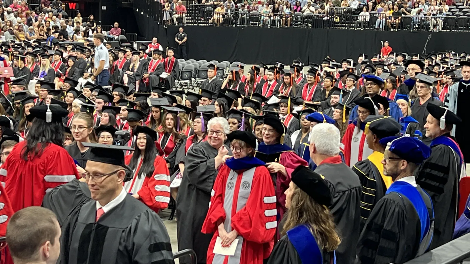hooding ceremony at the Schottenstein Center. Rows of graduating students in scarlet and grey regalia are seated. Graduate school staff help professors place hoods over the heads of their students.